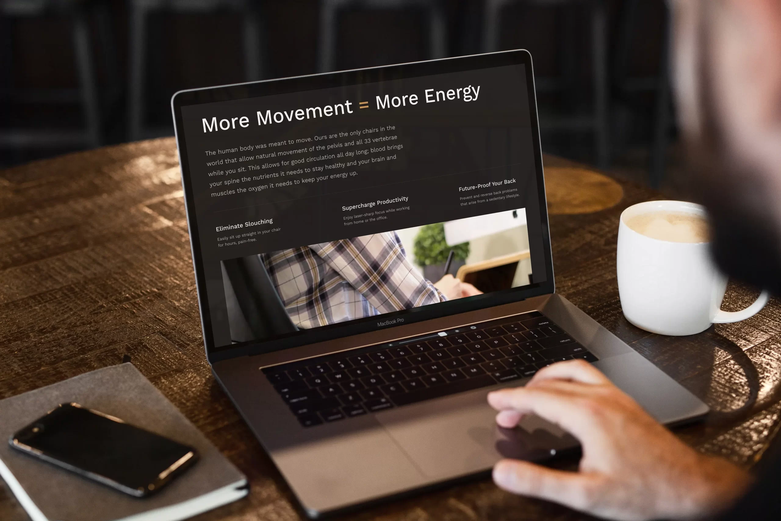 A laptop on a wooden café table shows the Axion website. The hero reads “More Movement = More Energy,” with a short paragraph and three benefit blocks: Eliminate Slouching, Supercharge Productivity, and Future-Proof Your Back. A video thumbnail sits below the copy. A white mug, notebook, and phone are beside the laptop; a hand rests near the trackpad.
